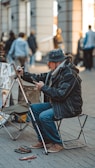 A street artist is sitting on a small chair, engrossed in drawing or painting on a canvas set on an easel. The artist is dressed in warm clothing, including a hat and a scarf. Various art supplies are scattered on the ground near the artist. In the background, several people are walking around, and the setting appears to be a bustling urban environment with buildings and shops.