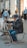 A street artist is sitting on a small chair, engrossed in drawing or painting on a canvas set on an easel. The artist is dressed in warm clothing, including a hat and a scarf. Various art supplies are scattered on the ground near the artist. In the background, several people are walking around, and the setting appears to be a bustling urban environment with buildings and shops.