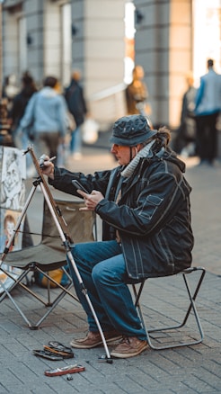 A street artist is sitting on a small chair, engrossed in drawing or painting on a canvas set on an easel. The artist is dressed in warm clothing, including a hat and a scarf. Various art supplies are scattered on the ground near the artist. In the background, several people are walking around, and the setting appears to be a bustling urban environment with buildings and shops.