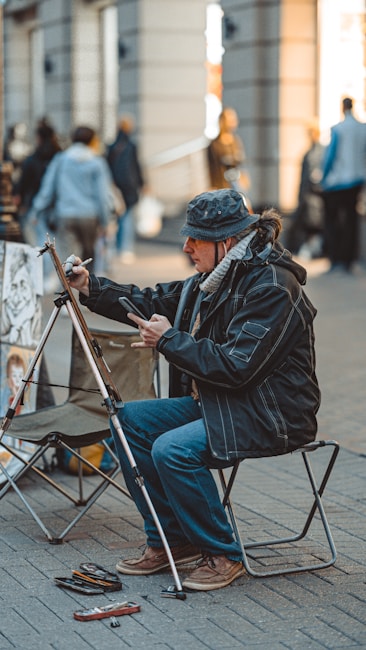 A street artist is sitting on a small chair, engrossed in drawing or painting on a canvas set on an easel. The artist is dressed in warm clothing, including a hat and a scarf. Various art supplies are scattered on the ground near the artist. In the background, several people are walking around, and the setting appears to be a bustling urban environment with buildings and shops.