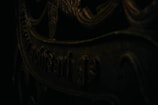 A close-up view of a dark, ornate metal surface with intricate patterns and partially visible text. The lighting casts shadows, enhancing the texture and giving a mysterious feel.