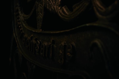 A close-up view of a dark, ornate metal surface with intricate patterns and partially visible text. The lighting casts shadows, enhancing the texture and giving a mysterious feel.