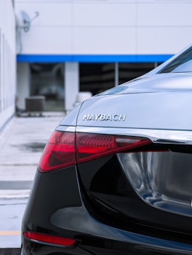 The rear of a luxury Maybach car with sleek black finish and distinct chrome accents. The taillight is illuminated with bright red LEDs. The car is parked in a modern, urban setting with a building in the background.