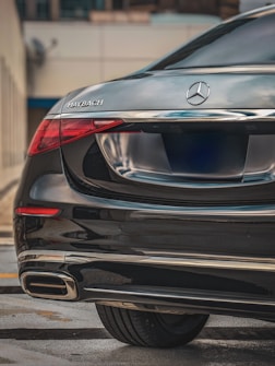 A luxury vehicle featuring the rear portion of a Mercedes-Maybach. The car has a sleek black finish with chrome accents, prominently displaying the Maybach logo on the left side and the Mercedes emblem in the center. The taillight design is modern and eye-catching, reflecting the high-end nature of the vehicle.