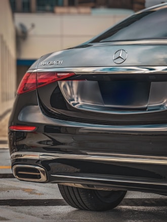 A luxury vehicle featuring the rear portion of a Mercedes-Maybach. The car has a sleek black finish with chrome accents, prominently displaying the Maybach logo on the left side and the Mercedes emblem in the center. The taillight design is modern and eye-catching, reflecting the high-end nature of the vehicle.
