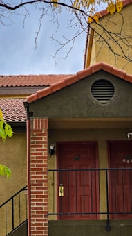 A building facade with a tiled roof and two red doors, featuring a small brick column. The structure includes a black metal railing and a vent above the doors. Branches with a few yellow leaves hang in the foreground.