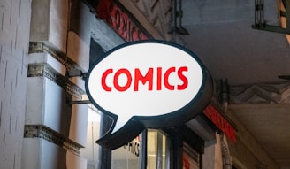 A brightly lit comic store sign shaped like a speech bubble with the word 'COMICS' written in bold red letters. The sign contrasts against the night sky and exterior of the building, suggesting an urban setting.