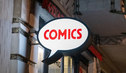 A brightly lit comic store sign shaped like a speech bubble with the word 'COMICS' written in bold red letters. The sign contrasts against the night sky and exterior of the building, suggesting an urban setting.