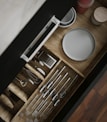 A wooden kitchen drawer is open, revealing neatly organized utensils including spoons, forks, and a whisk. Adjacent to the utensils, plates and a small container with red contents are visible. The drawer is displayed with a sleek handle, and the organization is visually appealing.
