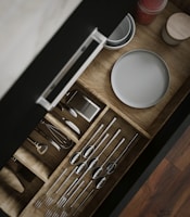 A wooden kitchen drawer is open, revealing neatly organized utensils including spoons, forks, and a whisk. Adjacent to the utensils, plates and a small container with red contents are visible. The drawer is displayed with a sleek handle, and the organization is visually appealing.