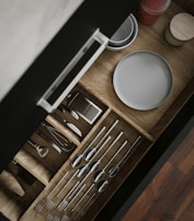 A wooden kitchen drawer is open, revealing neatly organized utensils including spoons, forks, and a whisk. Adjacent to the utensils, plates and a small container with red contents are visible. The drawer is displayed with a sleek handle, and the organization is visually appealing.
