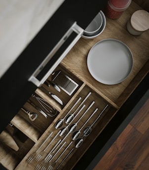 A wooden kitchen drawer is open, revealing neatly organized utensils including spoons, forks, and a whisk. Adjacent to the utensils, plates and a small container with red contents are visible. The drawer is displayed with a sleek handle, and the organization is visually appealing.