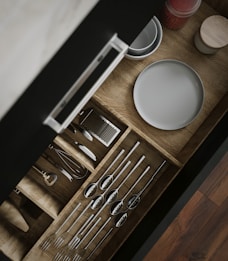A wooden kitchen drawer is open, revealing neatly organized utensils including spoons, forks, and a whisk. Adjacent to the utensils, plates and a small container with red contents are visible. The drawer is displayed with a sleek handle, and the organization is visually appealing.