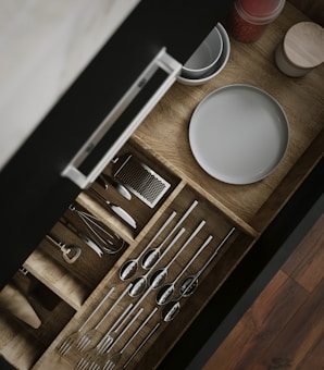 A wooden kitchen drawer is open, revealing neatly organized utensils including spoons, forks, and a whisk. Adjacent to the utensils, plates and a small container with red contents are visible. The drawer is displayed with a sleek handle, and the organization is visually appealing.