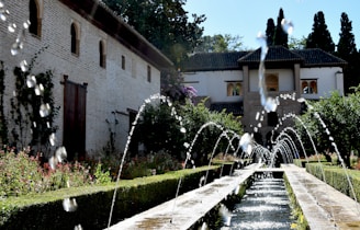 A beautifully landscaped garden features a series of arched water fountains cascading into a long rectangular pool. The garden is bordered by lush greenery and vibrant flowers. In the background, a historical building with arched windows and a rustic roof is visible. Tall trees provide a scenic backdrop, adding to the serene and peaceful ambiance.