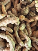 A collection of fresh turmeric roots, with their rough, brownish skin and vibrant orange interior peeking through in some spots. The roots are densely packed together, showcasing their natural, knobby form and earthy texture.