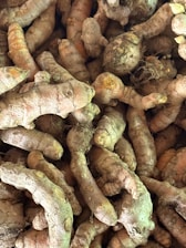 A collection of fresh turmeric roots, with their rough, brownish skin and vibrant orange interior peeking through in some spots. The roots are densely packed together, showcasing their natural, knobby form and earthy texture.