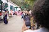 A lively outdoor gathering featuring a woman in a colorful dress speaking into a microphone. A group of people, some dressed in matching pink and white attire, stand nearby watching. The setting is festive with green banners and plants around a white building with blue shutters. A person in the foreground captures the scene on a smartphone.