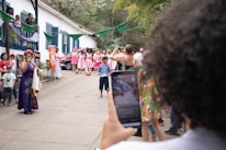 A lively outdoor gathering featuring a woman in a colorful dress speaking into a microphone. A group of people, some dressed in matching pink and white attire, stand nearby watching. The setting is festive with green banners and plants around a white building with blue shutters. A person in the foreground captures the scene on a smartphone.