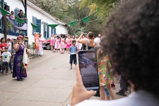 A lively outdoor gathering featuring a woman in a colorful dress speaking into a microphone. A group of people, some dressed in matching pink and white attire, stand nearby watching. The setting is festive with green banners and plants around a white building with blue shutters. A person in the foreground captures the scene on a smartphone.