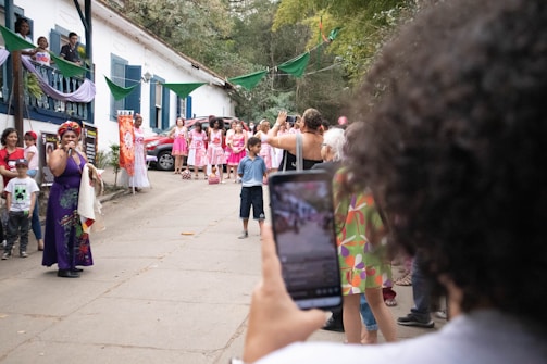 A lively outdoor gathering featuring a woman in a colorful dress speaking into a microphone. A group of people, some dressed in matching pink and white attire, stand nearby watching. The setting is festive with green banners and plants around a white building with blue shutters. A person in the foreground captures the scene on a smartphone.