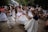 A group of people is dancing outdoors, wearing white dresses and colorful sashes. The scene is lively with many onlookers in casual attire in the background, suggesting a festive or cultural event. The setting appears to be a garden or park area with trees and some buildings nearby.