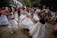 A group of people is dancing outdoors, wearing white dresses and colorful sashes. The scene is lively with many onlookers in casual attire in the background, suggesting a festive or cultural event. The setting appears to be a garden or park area with trees and some buildings nearby.