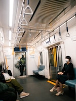 An interior view of a subway train car with several people seated. The car features empty aisle space and seats along the walls. Overhead, there are numerous safety handles for standing passengers. A person wearing a mask and dark clothing is seated on the right side. Another individual is seated on the left holding a phone, while a security personnel is visible standing near the far door.