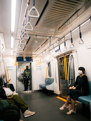 An interior view of a subway train car with several people seated. The car features empty aisle space and seats along the walls. Overhead, there are numerous safety handles for standing passengers. A person wearing a mask and dark clothing is seated on the right side. Another individual is seated on the left holding a phone, while a security personnel is visible standing near the far door.