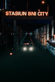 A train approaches a station at night, with bright headlights and a large illuminated sign above that reads 'STASIUN BNI CITY.' Two street lamps provide additional lighting, and the station is surrounded by city structures.