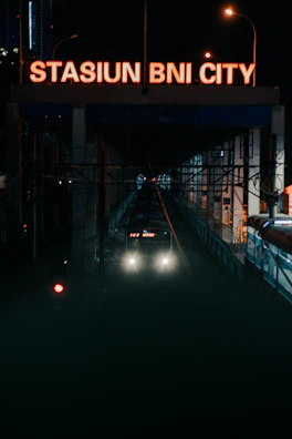 A train approaches a station at night, with bright headlights and a large illuminated sign above that reads 'STASIUN BNI CITY.' Two street lamps provide additional lighting, and the station is surrounded by city structures.