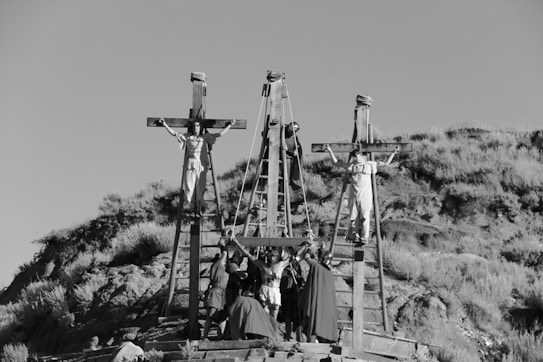 Three wooden crosses are erected on a hillside with figures attached to them, reminiscent of a crucifixion scene. Several people in period costumes are gathered at the base, involved in some activity. The landscape is dry with sparse vegetation.