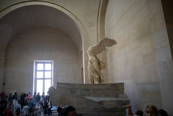 A large stone sculpture of a winged figure is prominently displayed in a spacious, arched hall. The sculpture stands on a pedestal, depicting a figure with wings spread, suggesting movement. Onlookers gather around, taking photos and admiring the artwork.