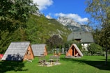 A serene landscape featuring small wooden A-frame cabins situated on a lush green lawn. The background showcases tall, rugged mountains with snow patches beneath a clear blue sky. Nearby trees add a sense of tranquility and nature.
