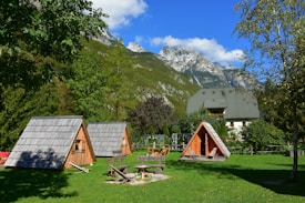 A serene landscape featuring small wooden A-frame cabins situated on a lush green lawn. The background showcases tall, rugged mountains with snow patches beneath a clear blue sky. Nearby trees add a sense of tranquility and nature.