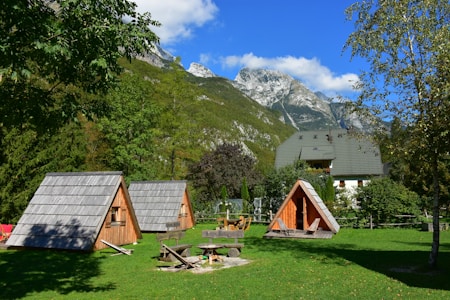 A serene landscape featuring small wooden A-frame cabins situated on a lush green lawn. The background showcases tall, rugged mountains with snow patches beneath a clear blue sky. Nearby trees add a sense of tranquility and nature.