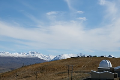 A landscape depicting snow-capped mountains under a blue sky with wispy clouds. In the foreground, rolling brown hills lead to an observatory with a domed roof and several parked cars nearby.