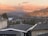 A scenic view of a hilly residential area with a backdrop of mountains and a lake. The houses are nestled among trees, and the mountains are softly illuminated by the setting sun, creating a warm, golden glow in the sky.