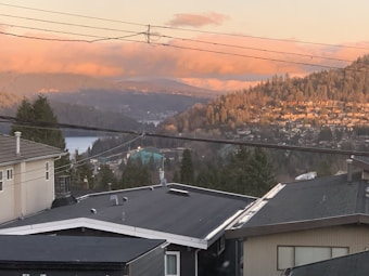 A scenic view of a hilly residential area with a backdrop of mountains and a lake. The houses are nestled among trees, and the mountains are softly illuminated by the setting sun, creating a warm, golden glow in the sky.