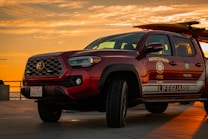 A Toyota lifeguard vehicle is parked on a concrete surface during a vibrant sunset. The vehicle is marked with emergency service logos, including 'Huntington Beach Fire' and 'Lifeguard'. The sky is filled with warm orange and yellow hues, suggesting a coastal setting.