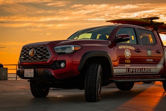 A Toyota lifeguard vehicle is parked on a concrete surface during a vibrant sunset. The vehicle is marked with emergency service logos, including 'Huntington Beach Fire' and 'Lifeguard'. The sky is filled with warm orange and yellow hues, suggesting a coastal setting.