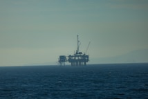 An offshore oil rig is situated in the sea, with cranes and structures visible against a hazy horizon. The water is dark and stretches out under a misty sky, creating a faint silhouette of distant land in the background.