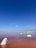 A serene landscape featuring a salt lake with calm, reddish-brown water under a clear blue sky. Large white salt formations protrude from the water, creating a striking contrast with the red hue. The horizon is distant, and the sky is vast, giving a sense of openness and tranquility.
