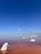 A serene landscape featuring a salt lake with calm, reddish-brown water under a clear blue sky. Large white salt formations protrude from the water, creating a striking contrast with the red hue. The horizon is distant, and the sky is vast, giving a sense of openness and tranquility.