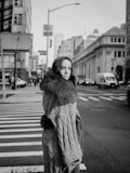 A woman wearing a textured coat with a fur collar stands on a city street. Tall buildings and urban architecture surround her, with a crosswalk and cars visible in the background. The image is captured in black and white, giving it a classic and timeless feel.