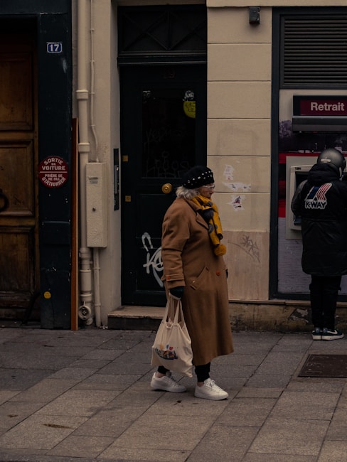 An elderly woman wearing a beige coat, a yellow-and-black scarf, a black hat, and white sneakers is walking on a sidewalk. She carries a shopping bag in her right hand. In the background, there is a dark green door with graffiti, and a person at an ATM machine dressed in a black jacket and a motorcycle helmet.