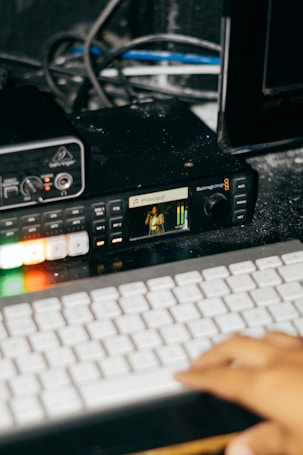 A close-up view of a keyboard and some audio or video equipment. A hand is visible, which suggests interaction with the keyboard. The equipment includes buttons that are illuminated and a small screen displaying an image and text with 'Principal' written on it.