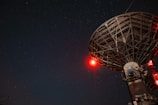 A large satellite dish under a starry night sky with a prominent red light illuminating part of the structure.