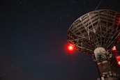 A large satellite dish under a starry night sky with a prominent red light illuminating part of the structure.