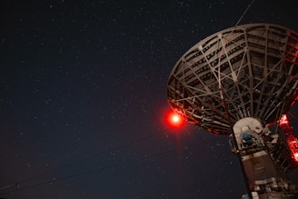 A large satellite dish under a starry night sky with a prominent red light illuminating part of the structure.